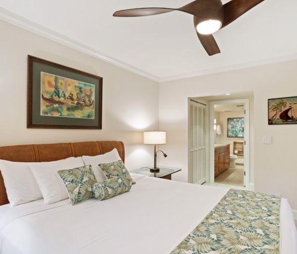 The image shows a cozy bedroom with a bed, decorative pillows, a ceiling fan, and artwork on the walls. It exudes a warm ambiance.