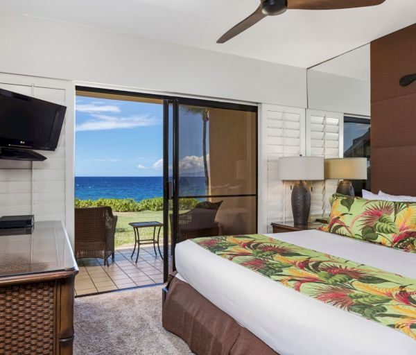 The image shows a stylish hotel room with a tropical-themed bedspread, a large window offering ocean views, and modern decor.