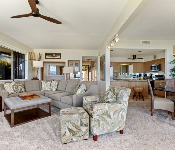 The image shows a cozy living room with a sectional sofa, tropical-patterned chairs, and an open kitchen in a bright setting.