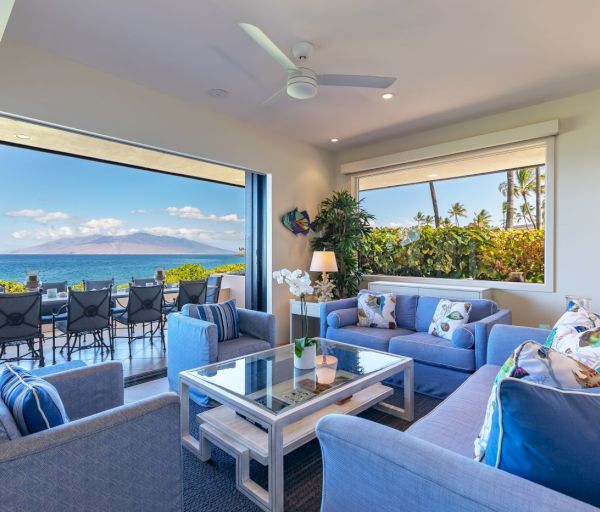The image shows a bright, airy living room with plush blue furniture and a view of the ocean through large windows.