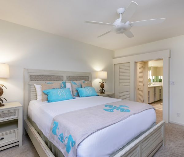 The image shows a cozy bedroom with a queen-sized bed, light decor, ceiling fan, and adjoining bathroom area. It's inviting and modern.