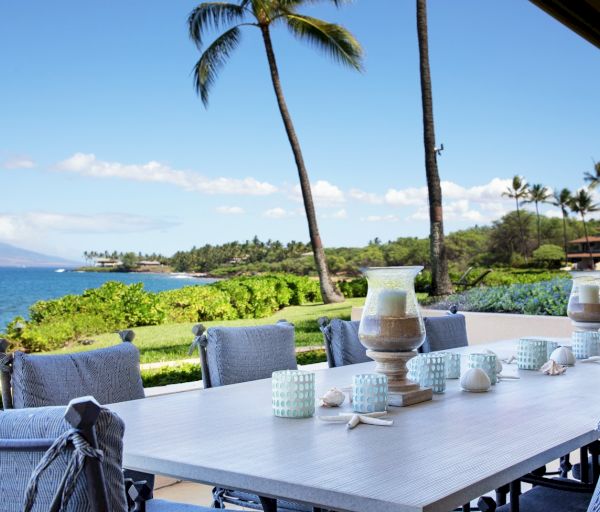 The image shows a serene patio with a dining table, overlooking a tropical landscape and ocean under a clear blue sky.