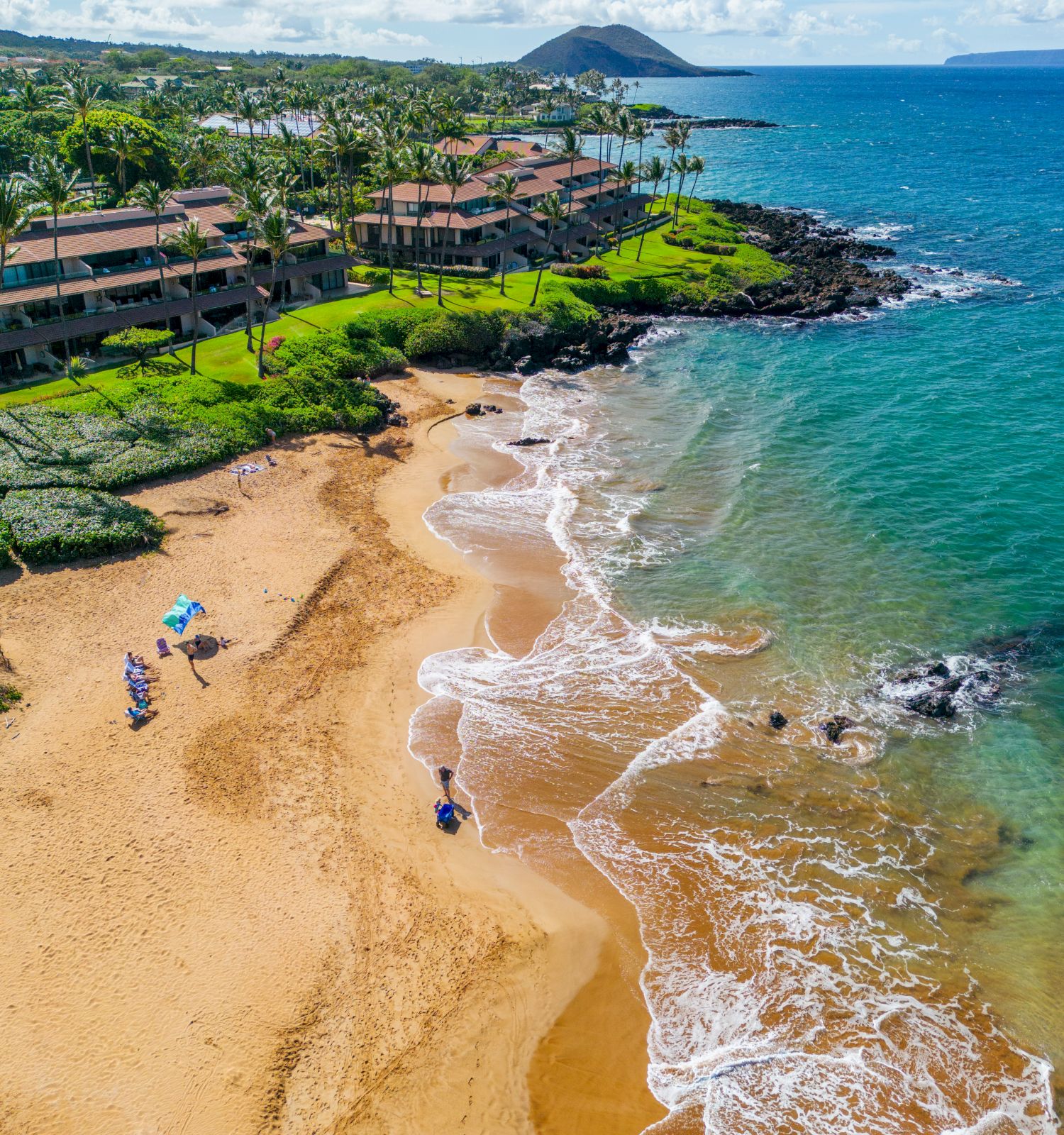 A scenic beach with golden sand, gentle waves, and green landscapes, bordered by buildings and a distant mountain.
