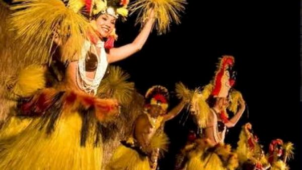 The image shows performers in vibrant yellow costumes, dancing with joyful expressions and traditional headdresses.