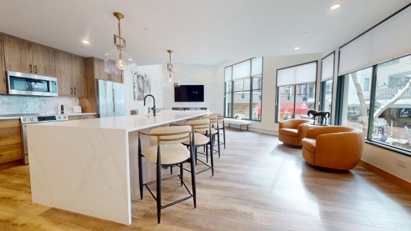 The image shows a modern kitchen with a large island, sleek appliances, and cozy seating, featuring large windows and natural light.