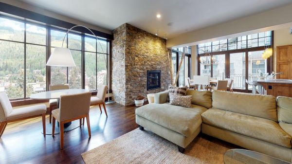 The image features a modern living area with a stone fireplace, large windows, a cozy couch, and a dining table nearby.