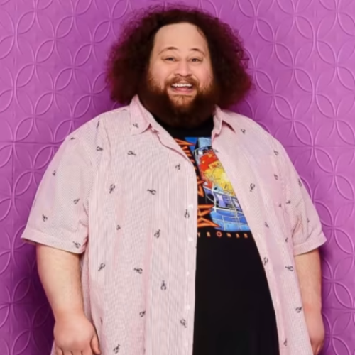 The image features a smiling man with curly hair wearing a patterned button-up shirt and a graphic t-shirt against a purple background.