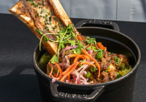 The image shows a pot containing meat, vegetables, and a large bone with herbs on top for garnish. It looks delicious and hearty.