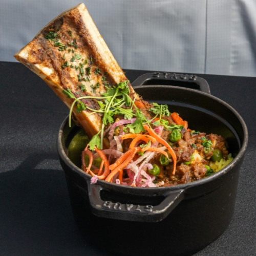 The image shows a pot containing meat, vegetables, and a large bone with herbs on top for garnish. It looks delicious and hearty.
