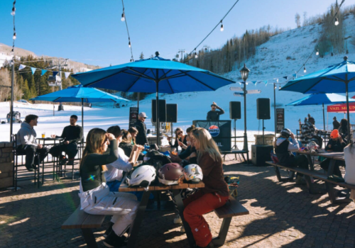 People enjoying a sunny day at a snowy outdoor caf&eacute;, seated under umbrellas, with a scenic mountain backdrop.