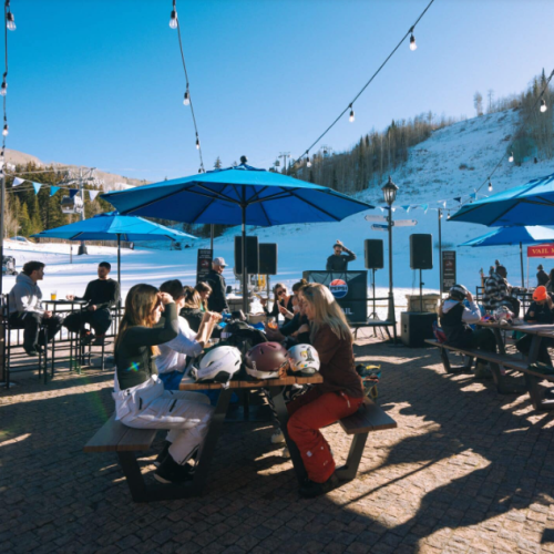 People enjoying a sunny day at a snowy outdoor caf&eacute;, seated under umbrellas, with a scenic mountain backdrop.