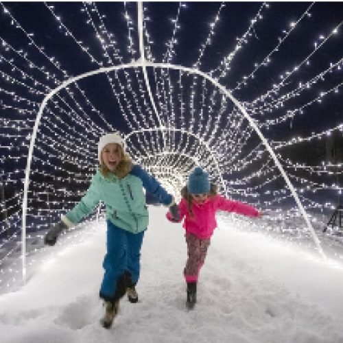Two children joyfully run through a snowy path illuminated by bright string lights in a magical winter setting.