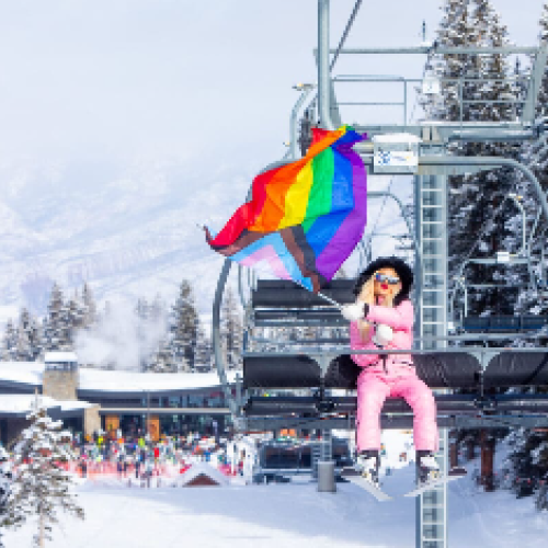 A person in pink ski gear is riding a chairlift, waving a rainbow flag amidst snowy mountains and skiers below.