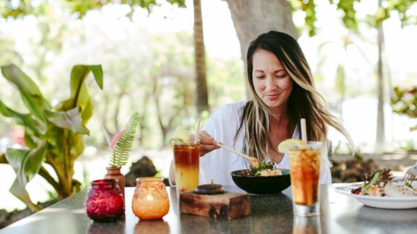 A woman is enjoying a meal outdoors, surrounded by drinks and candles, with lush greenery in the background. It's a peaceful setting.
