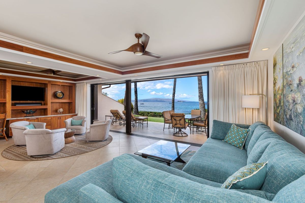 The image shows a stylish living room with a large window view, plush furniture, and a light, airy atmosphere.