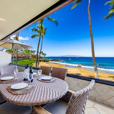 The image shows a sunny balcony with a dining table, palm trees, and a beautiful ocean view in the background. Perfect for relaxation.