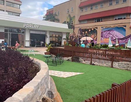 The image shows a landscaped area with a building, grassy spaces, and seating, set in a sunny urban environment.