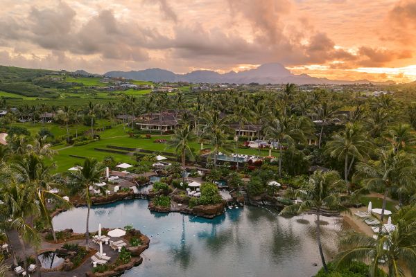 The image showcases a tropical resort with lush greenery, palm trees, and a serene water feature amidst a sunset backdrop.