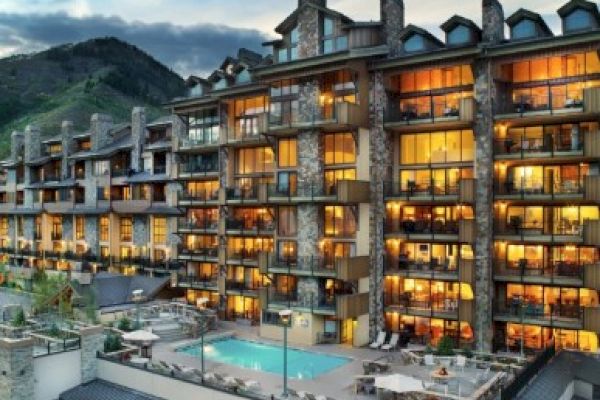 The image shows a modern hotel with large windows and a swimming pool, set against a mountain backdrop. It's inviting and scenic.