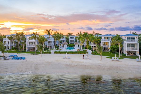 The image shows a serene beachfront property with modern houses, palm trees, and a colorful sunset sky. Relaxation awaits here.