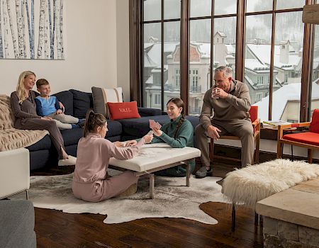 A cozy indoor scene with a family playing a game on a coffee table, while others relax on couches, with snowy views outside.