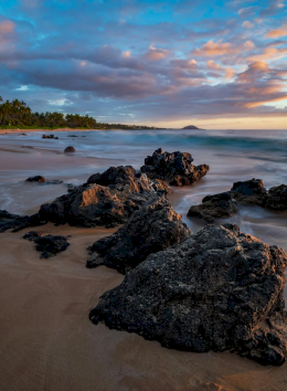 A tropical beach with rocky outcrops, gentle waves, palm trees in the distance, and a colorful sunset sky, serene and scenic.