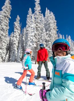 A group of four people skiing in a snowy landscape, surrounded by tall trees, under a clear blue sky. They look cheerful and bundled up.