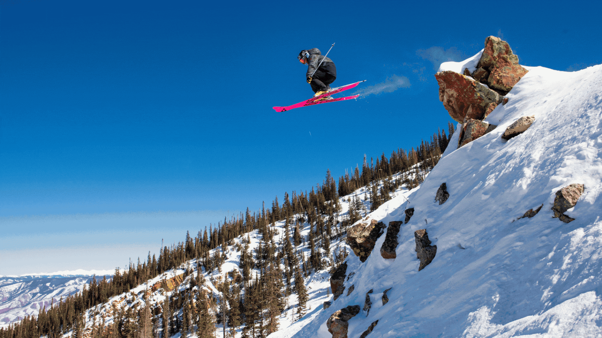 A skier with pink skis jumps off a snowy cliff against a clear blue sky and pine-covered mountains in the background.