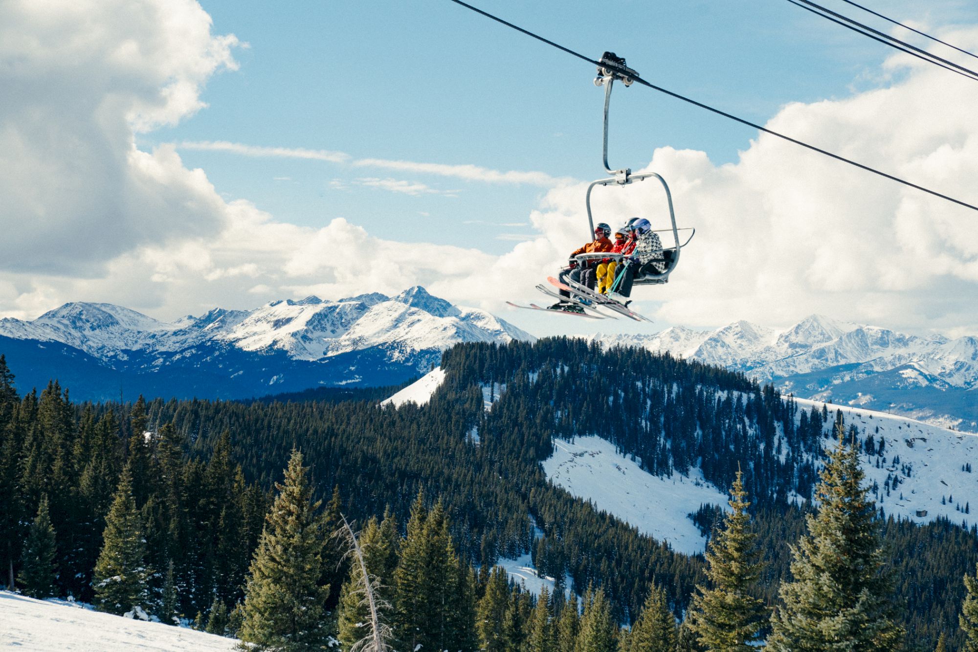 A ski lift carries people over snowy mountains, surrounded by evergreen trees and under a partly cloudy sky. It's a winter scene.