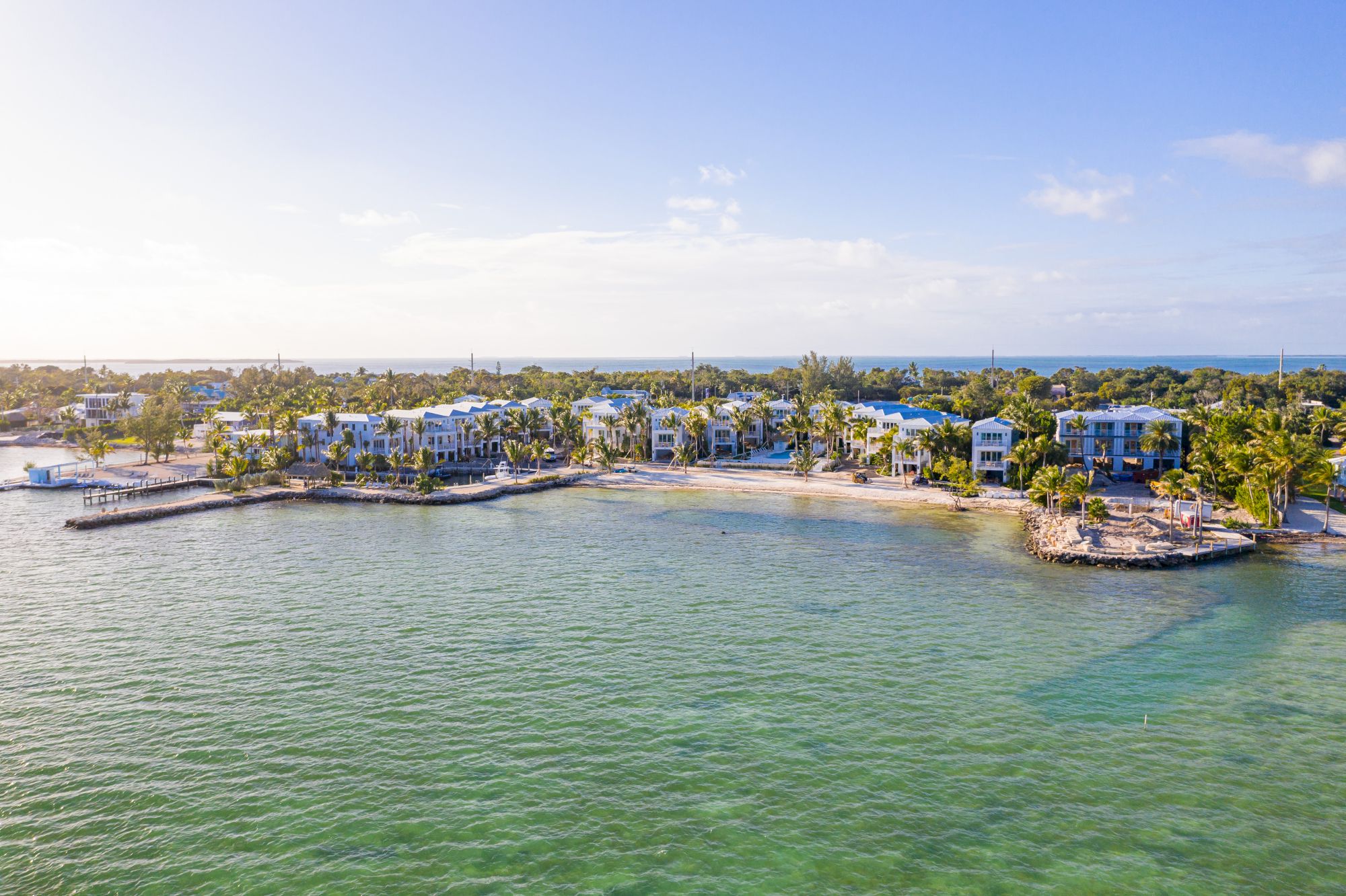 The image shows a tranquil coastal area with clear water, sandy shores, and modern buildings amidst lush greenery under a bright sky.
