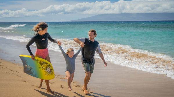 A family enjoys a day at the beach, with one adult holding a boogie board and walking with a child in the water.