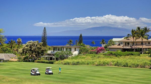 The image shows a lush golf course with two carts, a player, and beautiful ocean views with distant mountains. It&rsquo;s a scenic day.