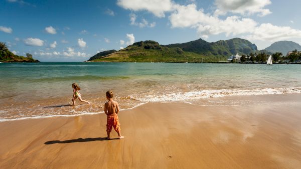 The image shows children playing on a sandy beach with calm water and mountains in the background under a blue sky.