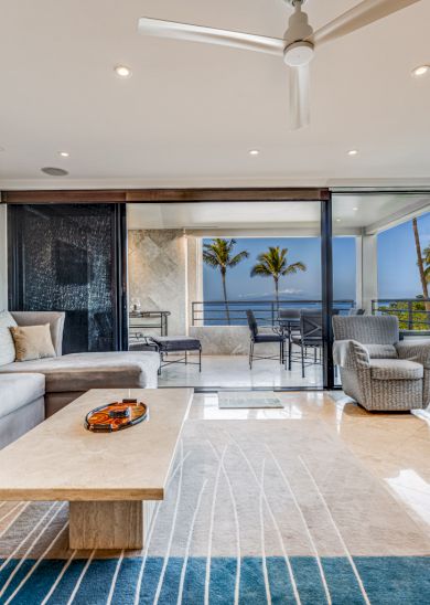 The image shows a modern living room with a large sofa, a coffee table, and stunning views of palm trees through glass doors.