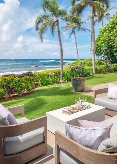 The image shows a serene patio with stylish seating, overlooking lush greenery and the ocean under a bright blue sky.