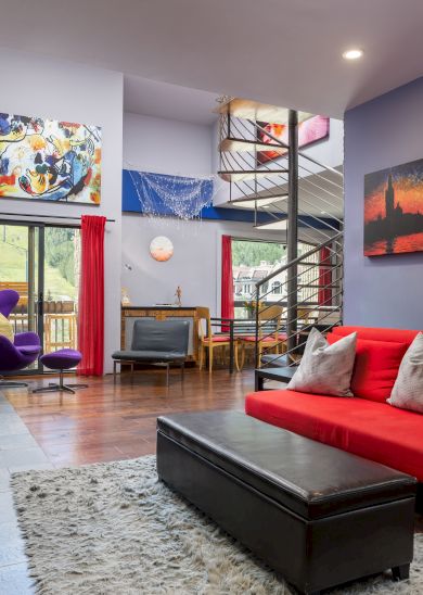 A modern living area with vibrant decor, featuring a red sofa, Buddha statue, spiral staircase, and colorful artwork on the walls.