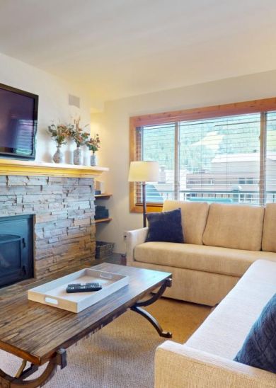 A cozy living room with a stone fireplace, TV, beige sofas, a wooden coffee table, and large window. Decor adds a warm touch to the space.