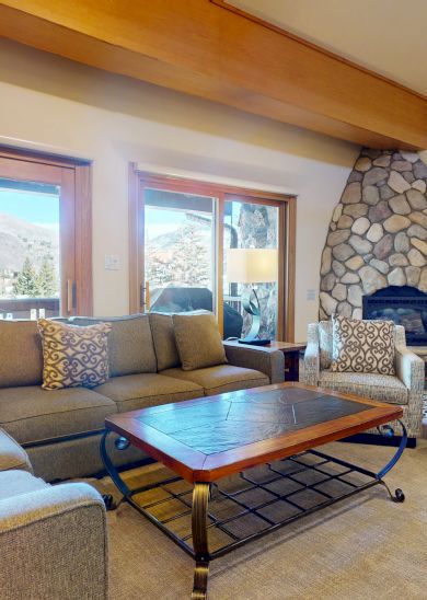 The image shows a cozy living room with a sectional sofa, stone fireplace, and large windows offering natural light.