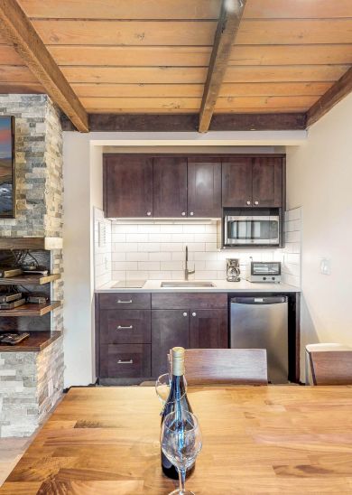 A cozy room with a fireplace, TV, kitchen, and dining table featuring a bottle of wine, under a wooden beamed ceiling with wall art.