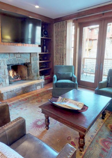 The image shows a cozy living room with a fireplace, bookshelves, comfortable seating, and large windows letting in natural light.