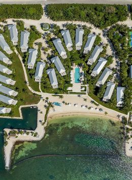 The image shows an aerial view of a coastal resort with bungalows, pools, and recreational areas by the water. It looks very inviting.