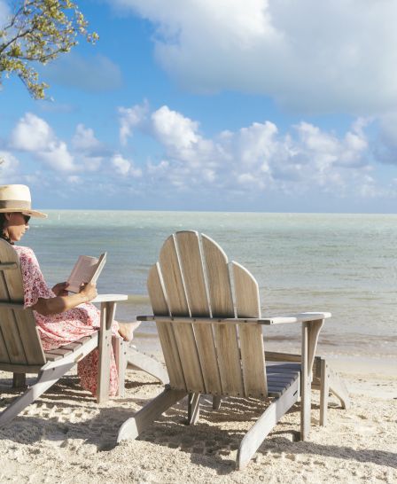 The image shows a person reading in a beach chair by the water, with a beautiful sky and gentle waves in the background.