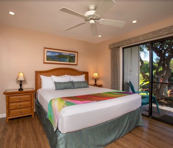 The image shows a cozy bedroom with a large bed, wooden furniture, and sliding glass doors opening to a patio with greenery.