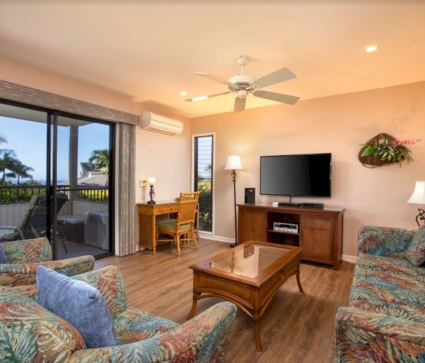 The image shows a cozy living room with colorful furniture, a desk, a TV, and large windows revealing a tropical view.