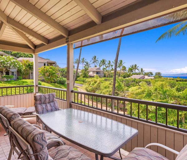 The image features a balcony with a table and chairs, overlooking lush greenery and a scenic ocean view in the distance.