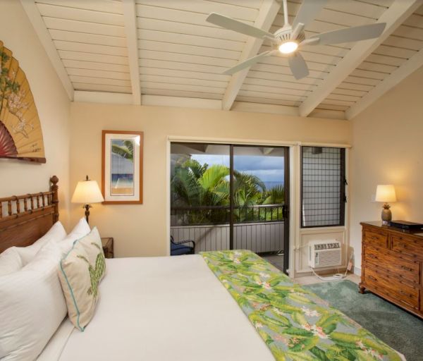 The image shows a cozy bedroom with a fan, a large bed, decorative pillows, and a view of greenery through the window.