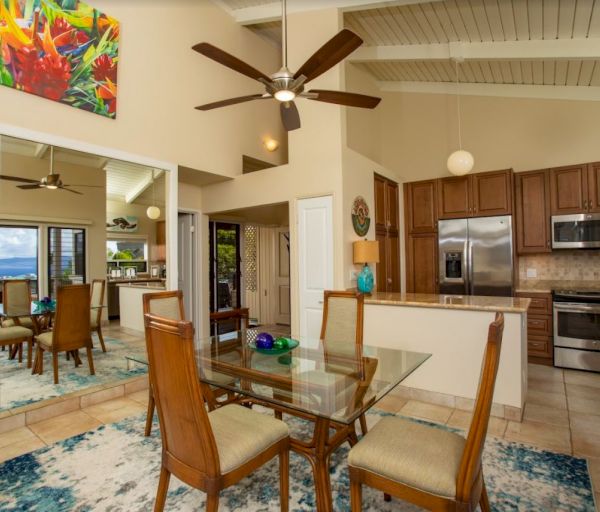 The image shows a modern open-concept living space with a dining area and kitchen, featuring large windows and tropical decor.