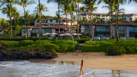 A person walks along a sandy beach with palm trees and a resort in the background, under a partly cloudy sky.