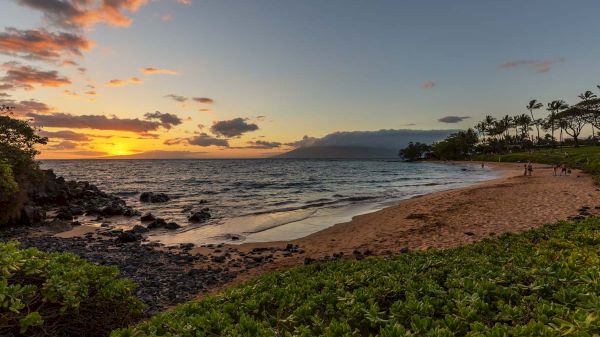 The image shows a serene beach at sunset, with gentle waves, distant mountains, and silhouetted palm trees against a colorful sky.