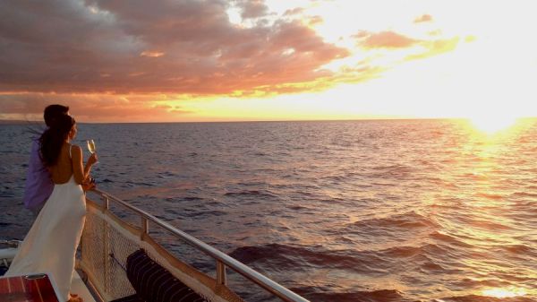A person in a white dress stands on a boat, watching a sunset over the ocean while holding a drink. It's a serene moment.